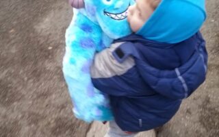 A young child in a blue hooded jacket walks outdoors, holding a large, blue plush toy resembling Sulley from Monsters, Inc. The ground is a mix of grey pavement and tarmac, suggesting a pavement or street. The child looks down at the toy affectionately.
