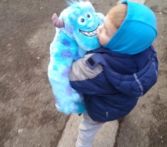 A young child in a blue hooded jacket walks outdoors, holding a large, blue plush toy resembling Sulley from Monsters, Inc. The ground is a mix of grey pavement and tarmac, suggesting a pavement or street. The child looks down at the toy affectionately.