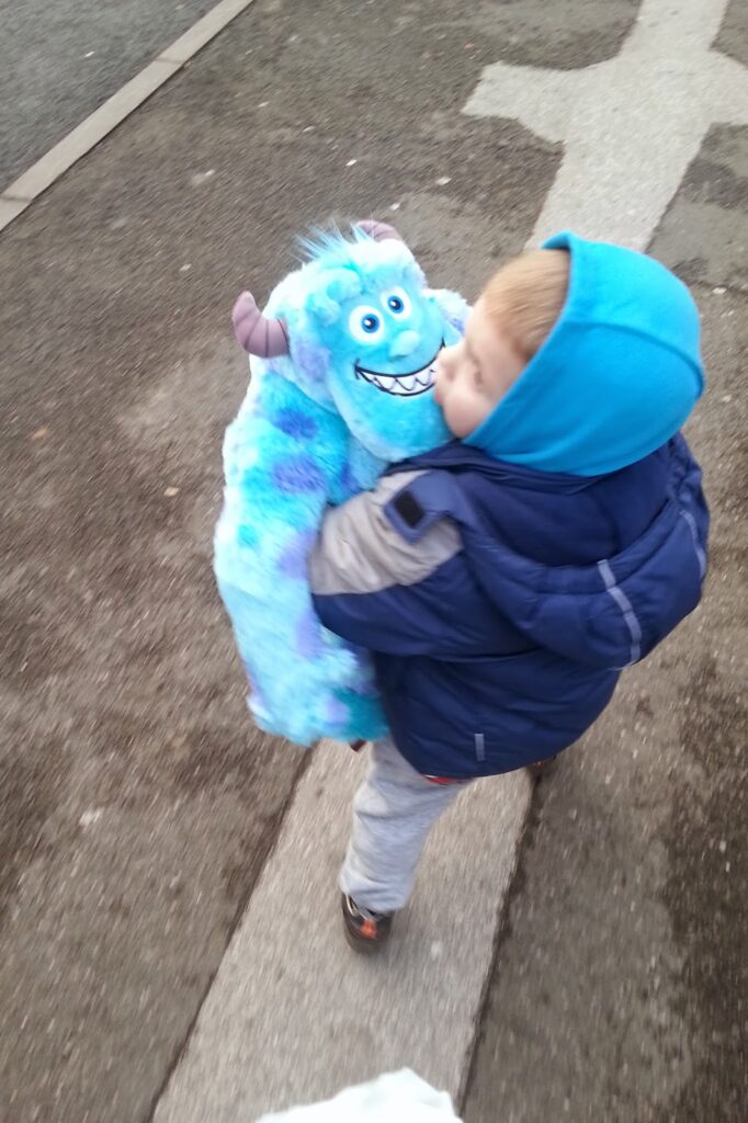 A young child in a blue hoodie and navy jacket walks outside on a pavement, hugging a large blue soft toy of Sulley from Monsters, Inc. The child looks happy and focused on the toy. The pavement has a white marking and some wet patches.