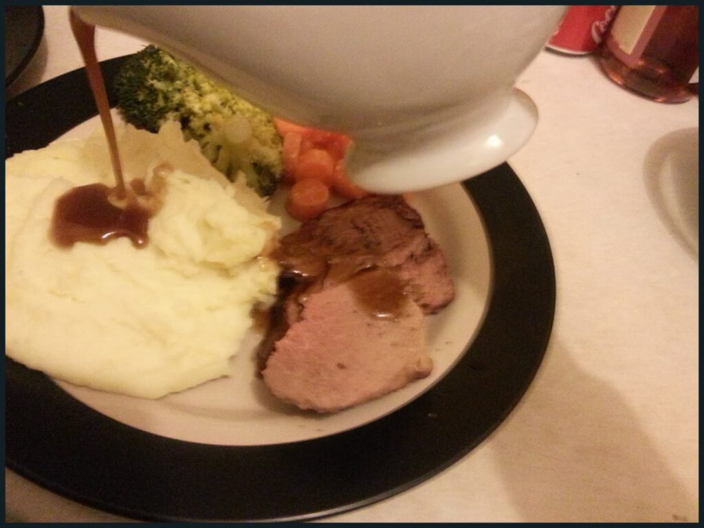 A hand pours brown gravy from a white gravy boat onto slices of roast beef, mashed potato, broccoli, and carrots on a round plate with a black rim, set on a light tablecloth.