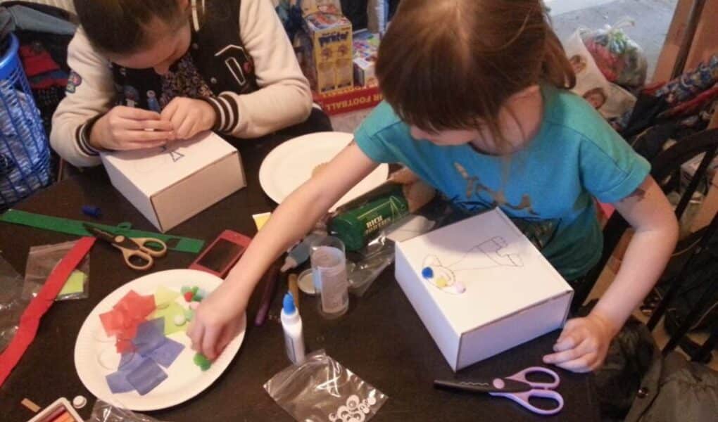 Two children sit at a table doing crafts. They decorate white boxes with glue, coloured tissue paper, foam shapes, and googly eyes. Scissors, felt-tip pens, and bottles of glue are scattered about, with craft materials on plates nearby.
