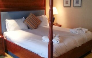 A neatly made four-poster bed with white linen and two towel sets sits in a warmly lit hotel room. There is a striped accent cushion, a bedside table with a lamp, framed art on the wall, and a yellow carpet with a subtle pattern.