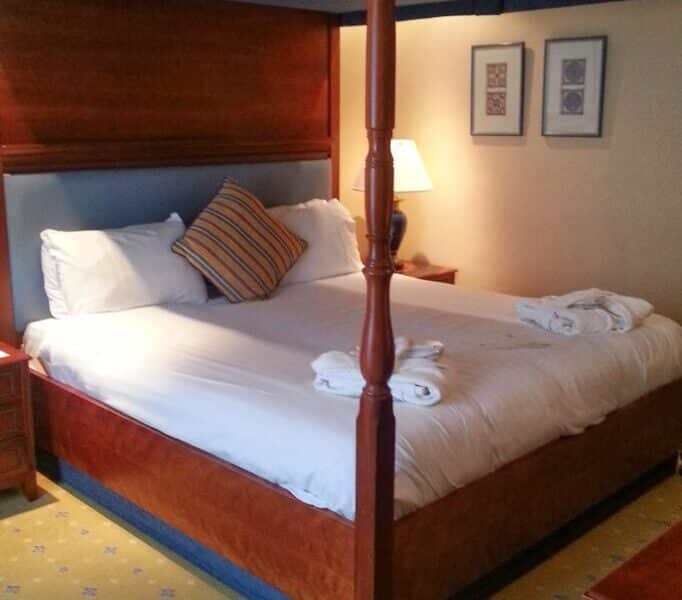 A neatly made four-poster bed with white linen and two towel sets sits in a warmly lit hotel room. There is a striped accent cushion, a bedside table with a lamp, framed art on the wall, and a yellow carpet with a subtle pattern.