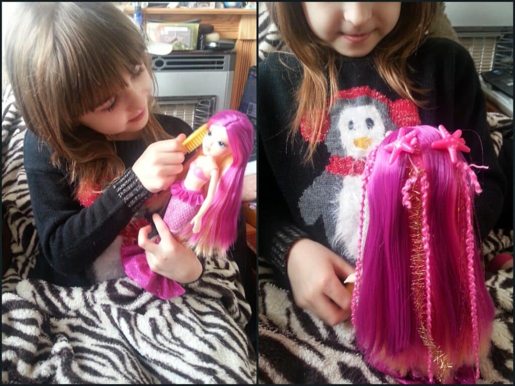 A young child with light brown hair, wearing a black jumper with a penguin design, sits on a zebra-print blanket. She is brushing the long pink hair of a mermaid doll. The doll’s hair is decorated with ribbons and golden strands.