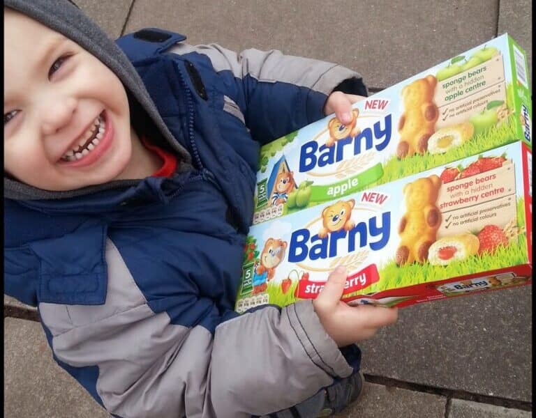 A smiling child in a blue and grey winter jacket holds two boxes of Barny sponge bear snacks, one apple flavour and one strawberry flavour, while standing on a paved surface outdoors. The child looks up at the camera, showing excitement.