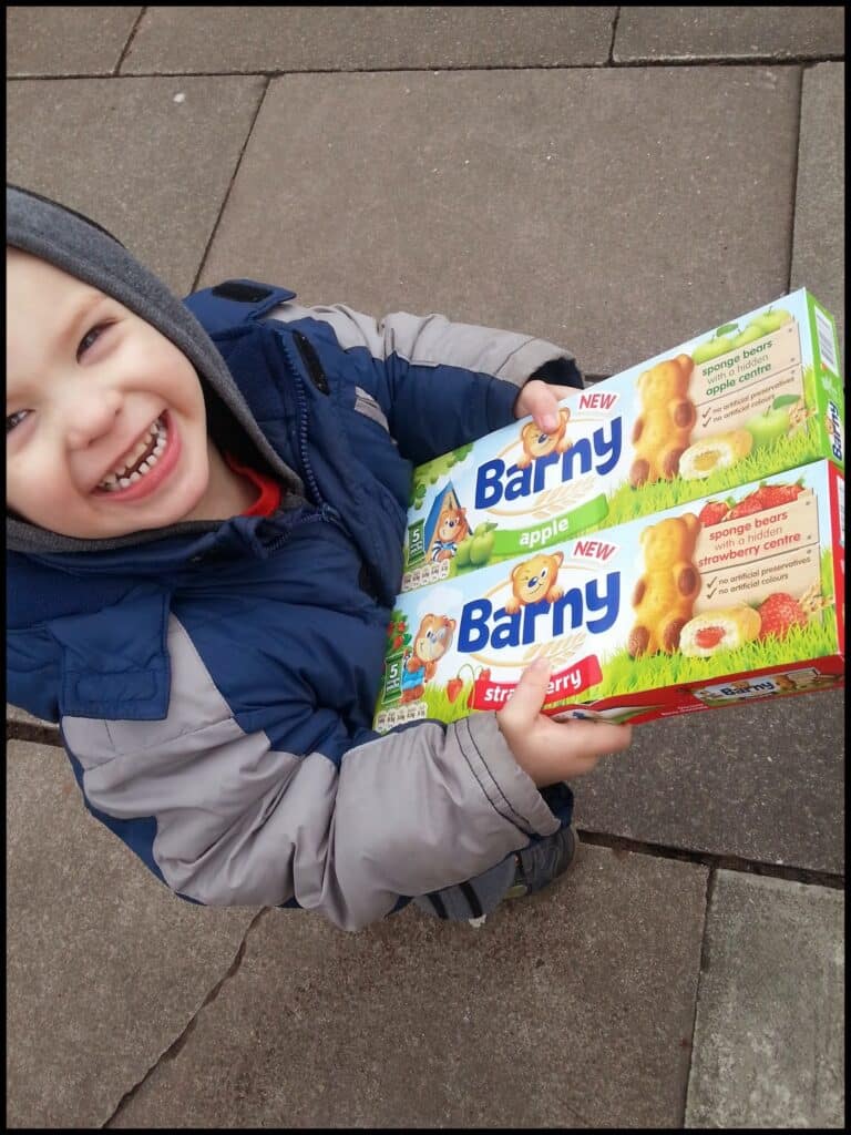 A smiling child in a grey and blue jacket holds two boxes of Barny cakes, one apple and one strawberry flavour, while standing on a pavement. The boxes display images of bear-shaped cakes with creamy centres and colourful branding.
