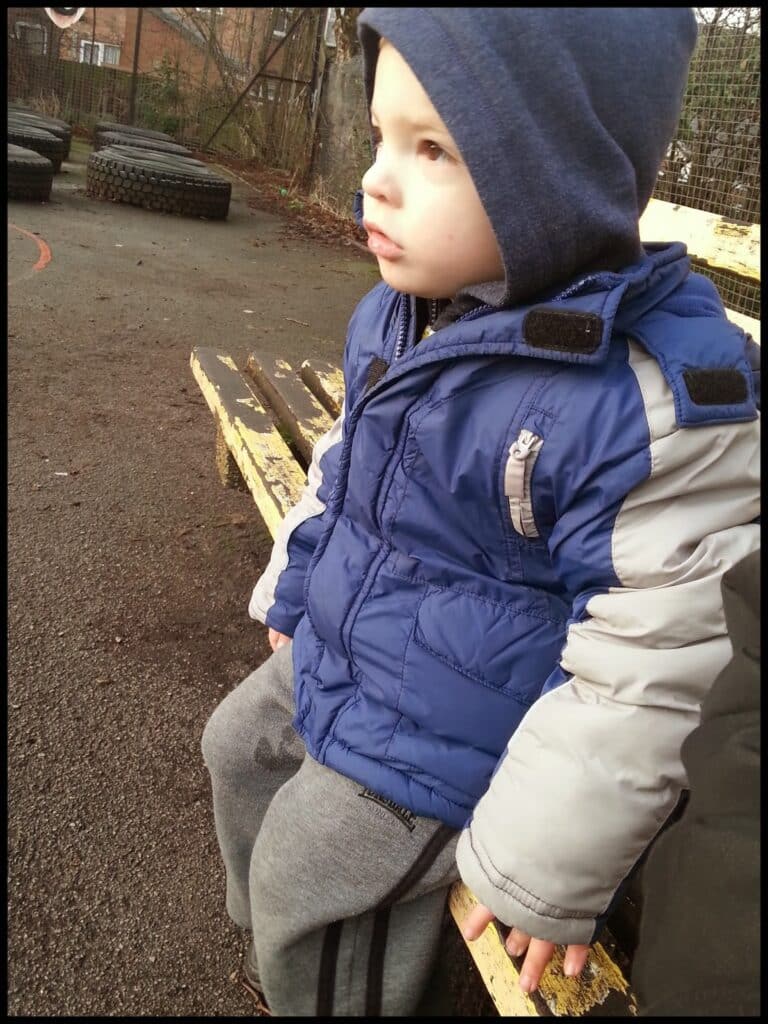 A young child wearing a blue and grey hooded winter coat and grey tracksuit bottoms sits on a worn yellow bench outdoors. The child looks thoughtfully into the distance; tyres and a wire fence are visible in the background, hinting at a playground or park setting.
