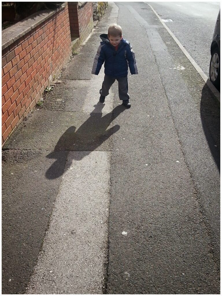 A young boy in a blue jacket and grey trousers stands on a pavement casting a large shadow. The sun is behind him, and the pavement runs alongside a red brick wall with a parked car partly visible on the right. The road stretches into the distance.