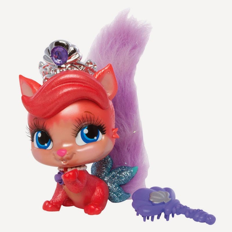 A toy squirrel with red fur, big blue eyes, long lashes, and a fluffy purple tail wears a silver tiara with a purple gem. It has blue glittery wings and a purple collar with a heart charm. A small purple brush shaped like a flower lies beside it.