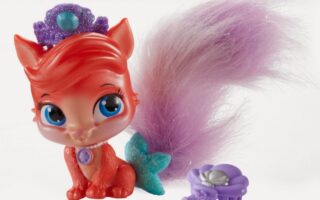 A red toy fox with large blue eyes, a purple jewelled tiara, and a fluffy purple tail sits beside a small, purple flower-shaped brush. The fox has a turquoise glittery bow on its tail and a purple gem necklace on its chest. The background is white.