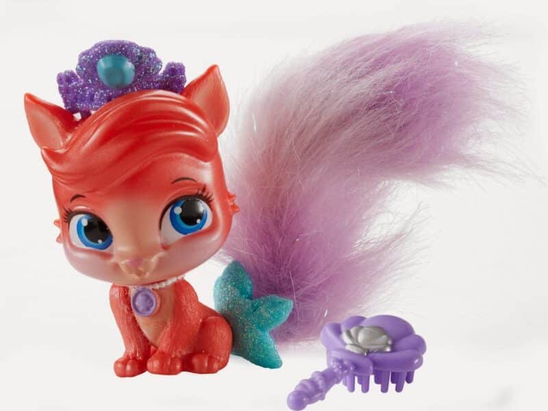 A red toy fox with large blue eyes, a purple jewelled tiara, and a fluffy purple tail sits beside a small, purple flower-shaped brush. The fox has a turquoise glittery bow on its tail and a purple gem necklace on its chest. The background is white.