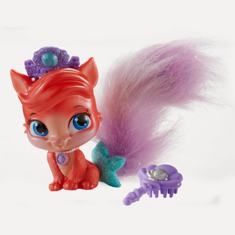 A red toy fox with large blue eyes, a purple jewelled tiara, and a fluffy purple tail sits beside a small, purple flower-shaped brush. The fox has a turquoise glittery bow on its tail and a purple gem necklace on its chest. The background is white.