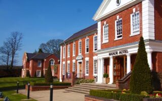 A large red-brick building with white columns and trim, labelled "Manor Hotel" above the entrance. Manicured lawns, hedges, and cone-shaped bushes line the walkway. Blue sky and leafless trees are visible in the background.