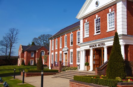 A large red-brick building with white columns and trim, labelled "Manor Hotel" above the entrance. Manicured lawns, hedges, and cone-shaped bushes line the walkway. Blue sky and leafless trees are visible in the background.