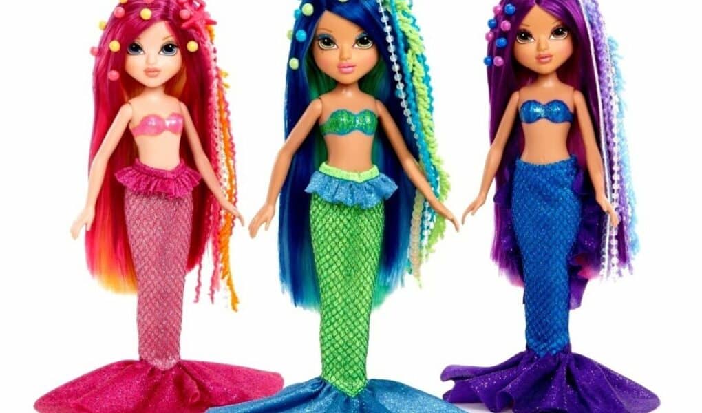 Three colourful mermaid dolls stand side by side. Each has long, vibrant hair adorned with beads and sea-themed accessories, and wears a sparkly bra top with a matching mermaid tail in pink, blue, and purple colours, respectively.
