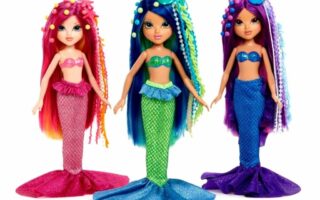 Three colourful mermaid dolls stand side by side. Each has long, vibrant hair adorned with beads and sea-themed accessories, and wears a sparkly bra top with a matching mermaid tail in pink, blue, and purple colours, respectively.