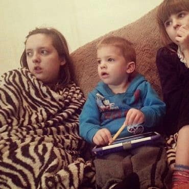 Three children sit closely on a sofa. The left child is wrapped in a zebra-striped blanket, the middle child holds a tablet and looks ahead, and the right child has a fringe, holding her hand to her mouth. All appear absorbed in what they are watching.