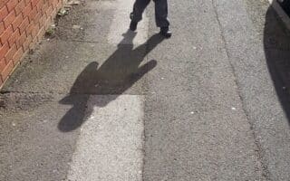 A young child in a blue jacket and grey trousers walks on a sunlit pavement, casting a prominent shadow that resembles the silhouette of a bear. A brick wall runs along the left side, and a parked car is visible on the right.