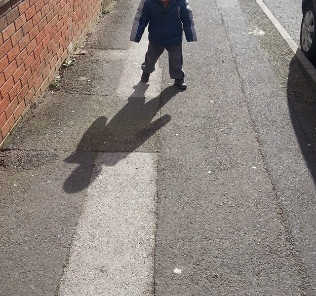 A young child in a blue jacket and grey trousers walks on a sunlit pavement, casting a prominent shadow that resembles the silhouette of a bear. A brick wall runs along the left side, and a parked car is visible on the right.