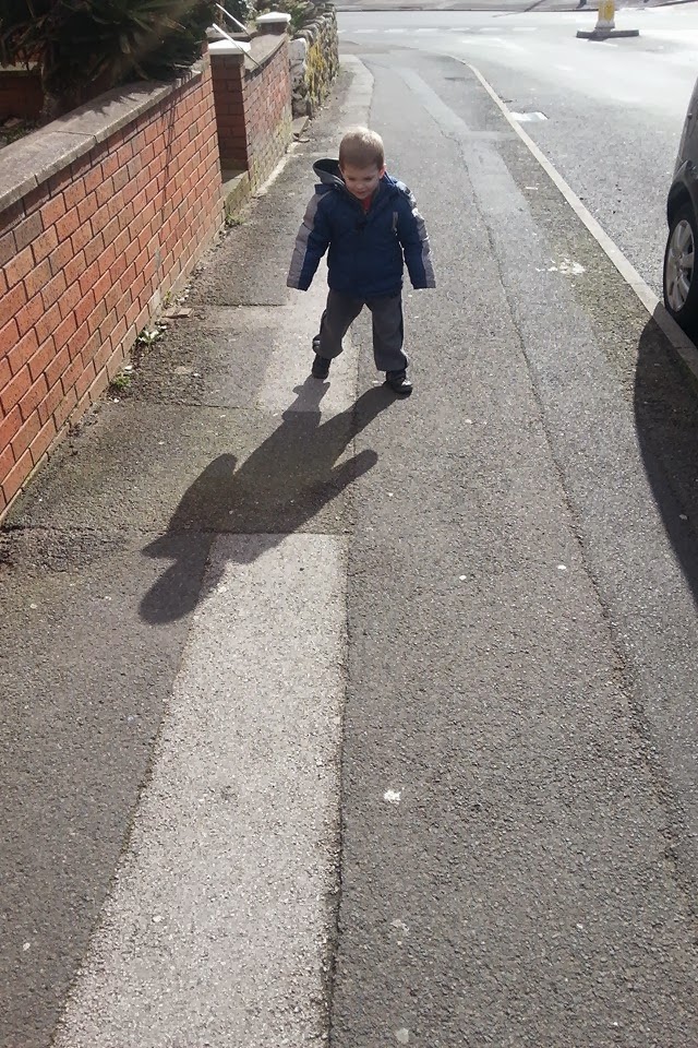 A young child in a blue jacket and grey trousers walks on a sunlit pavement, casting a prominent shadow that resembles the silhouette of a bear. A brick wall runs along the left side, and a parked car is visible on the right.