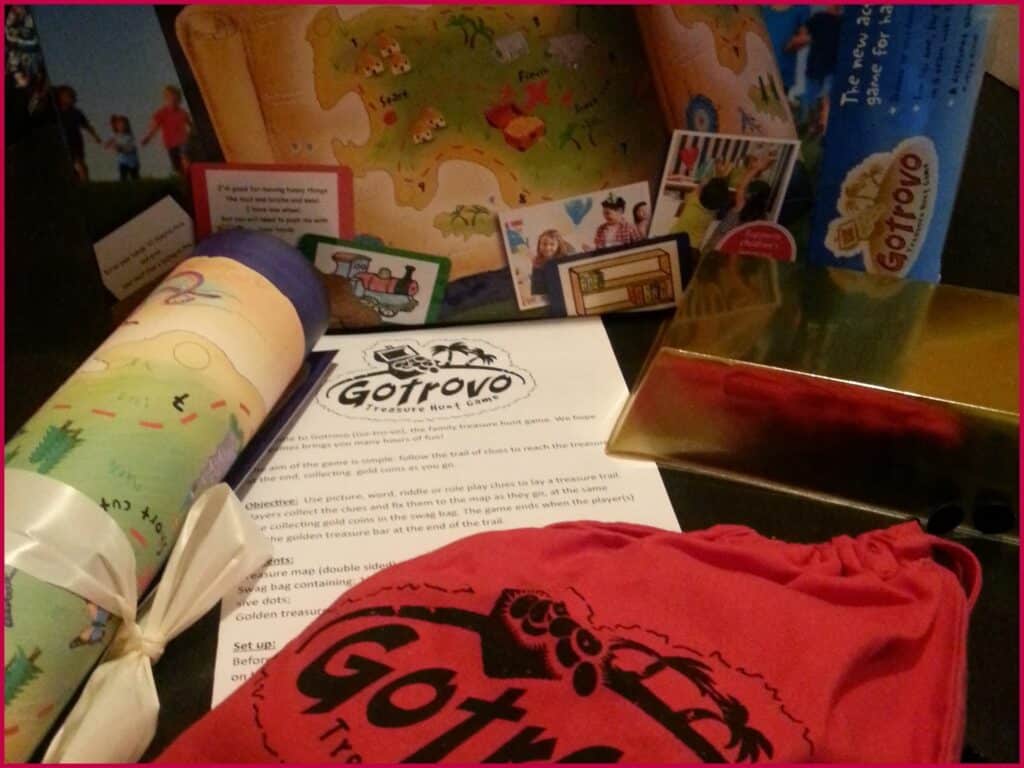 A Gotrovo Treasure Hunt kit is displayed, including a rolled-up playmat tied with ribbon, a red drawstring bag, a gold treasure box, game cards, instructions, and a treasure map, all arranged closely on a dark surface.