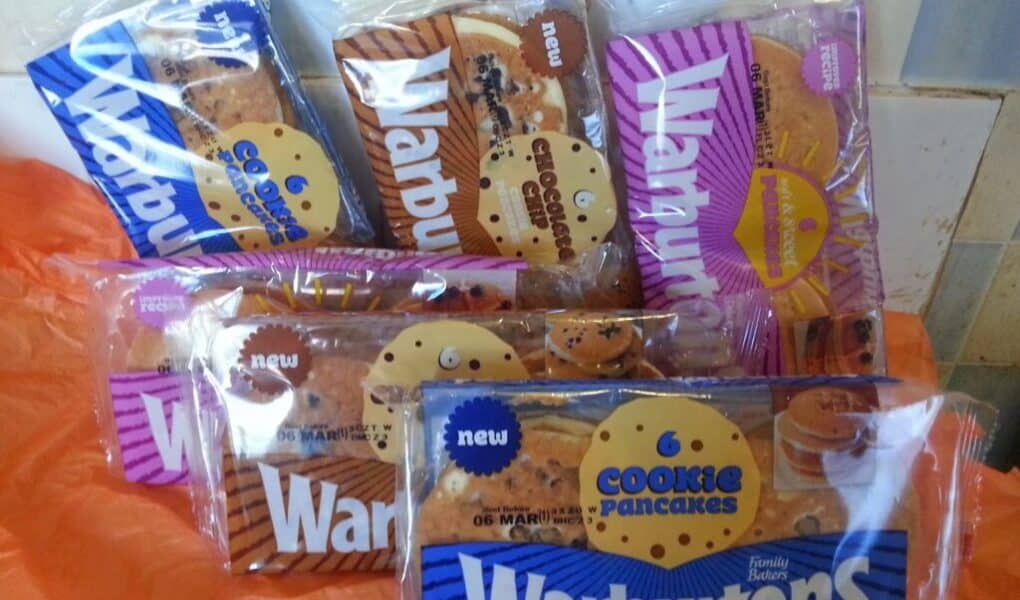 Several packs of Warburtons Cookie Pancakes are displayed on an orange surface. The packaging is clear with blue and white labels, showing the round pancakes inside. Some packs are stacked, and the background includes tiled walls.