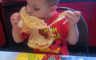 A young boy sits at a table, holding and biting into a large crêpe with both hands. He wears a red shirt with yellow lettering and is focused on eating. In front of him is a colourful plate with a floral pattern. The background shows a red kitchen.