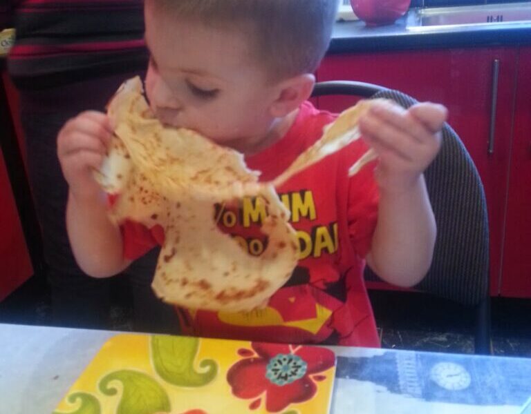 A young boy sits at a table, holding and biting into a large crêpe with both hands. He wears a red shirt with yellow lettering and is focused on eating. In front of him is a colourful plate with a floral pattern. The background shows a red kitchen.