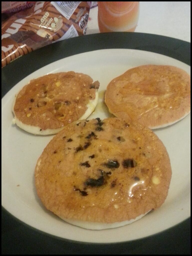 Three pancakes with visible chocolate chips and syrup are arranged on a white plate with a black rim. In the background, there is a partially visible bag of bread and a bottle, possibly containing juice or syrup.