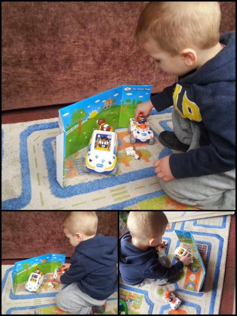 A young child with short blond hair, wearing a navy hoodie and grey trousers, kneels on a colourful carpet, playing with toy vehicles and a Paw Patrol-themed backdrop. He moves a white and orange toy car near a stationary white police car.