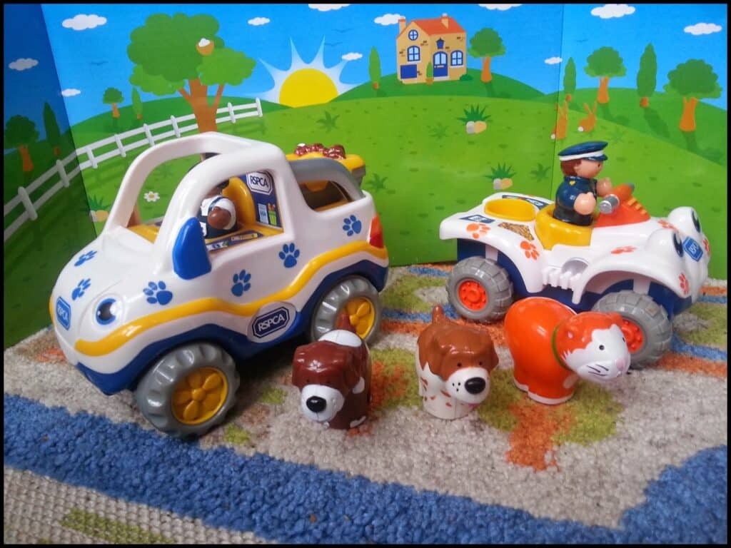 Two toy vehicles and four animal figurines are on a blue and green carpet. The left vehicle is white with blue paw prints and an animal driver. The right one has a person in uniform driving. The animals include two dogs, a cow, and an orange-striped tiger.