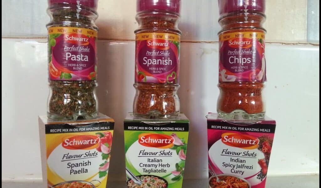 Three Schwartz spice blends in clear bottles with red lids are displayed on top of their matching Flavour Shots boxes: Spanish Pasta, Chips, and Chicken. The boxes below them are Spanish Paella, Creamy Herb Tagliatelle, and Spicy Jalfrezi Curry.
