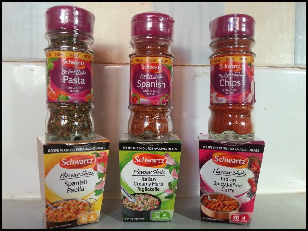 Three Schwartz spice blends in tall plastic bottles are shown, each placed atop a matching box of Flavour Shots. Flavours are Spanish Pasta, Chips, and Spanish Paella (left); Italian Creamy Herb Tagliatelle (centre); Indian Spicy Jalfrezi Curry (right).