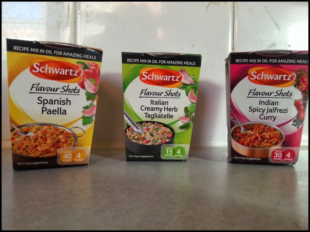 Three Schwartz Flavour Shots boxes are lined up on a worktop. Flavours are Spanish Paella, Italian Creamy Herb Tagliatelle, and Indian Spicy Jalfrezi Curry. Each box displays a bowl of the dish and serving size information, against a tiled wall.