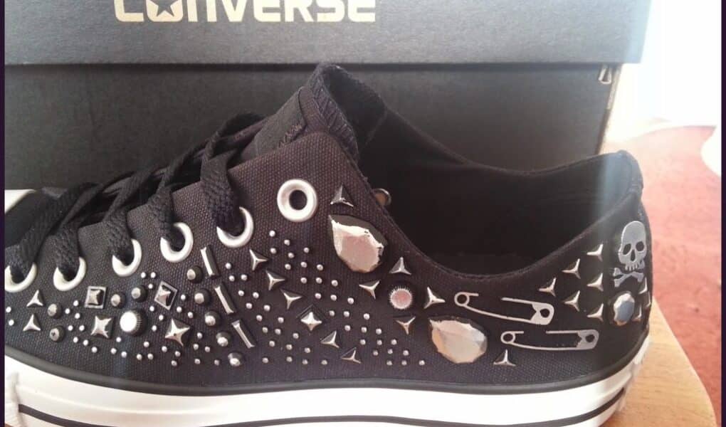 A black Converse trainer decorated with silver studs, safety pins, round gems, and a small skull graphic is shown in front of a black Converse shoe box. The shoe has white rubber soles and black laces, and is displayed on a wooden surface.