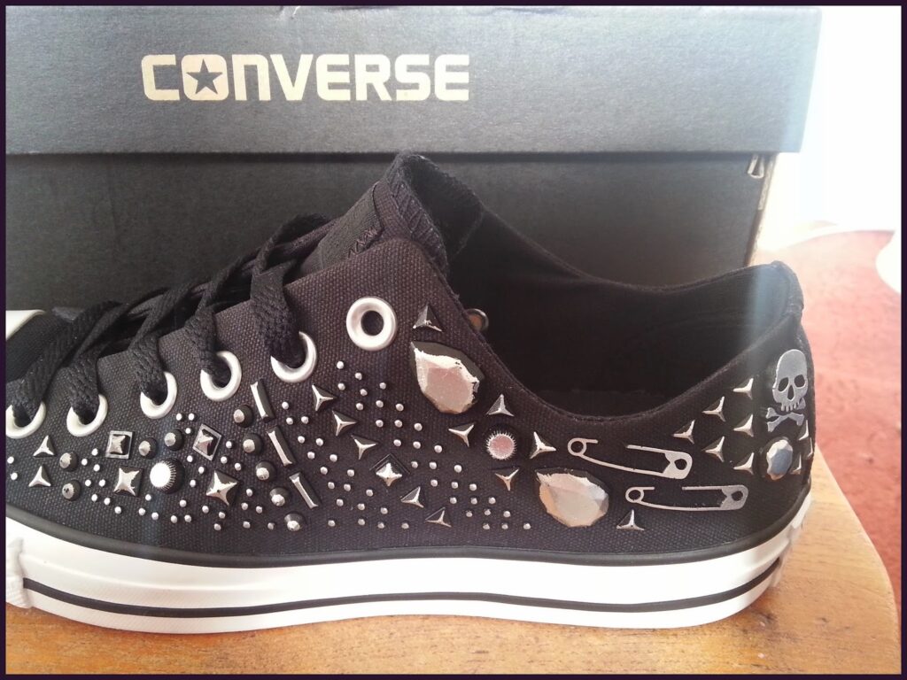 A black Converse trainer decorated with silver studs, spikes, safety pins, and a small skull emblem. The shoe is placed in front of a black Converse shoe box, and the shoe’s white sole features a black stripe. The background includes a wooden surface and a carpet.