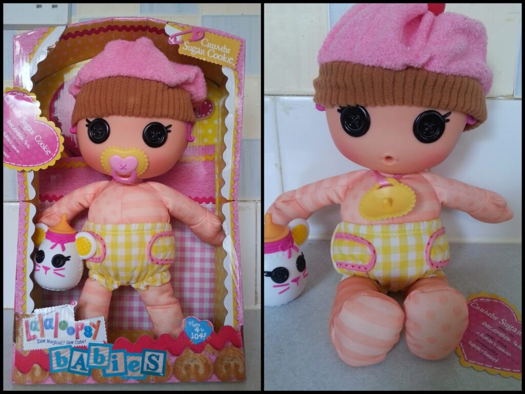 A soft pink doll with large black button eyes, a yellow dummy, a pink and brown cupcake hat, yellow chequered shorts, and orange-striped legs is shown in its box on the left and out of the box on the right, holding a white pet with pink accents.