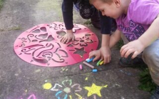 Two young children crouch on a paved path, using coloured chalk and a large red stencil to draw vibrant shapes and flowers on the ground. Colourful chalk drawings and patterns fill the pavement around their hands, with grass visible at the edges.