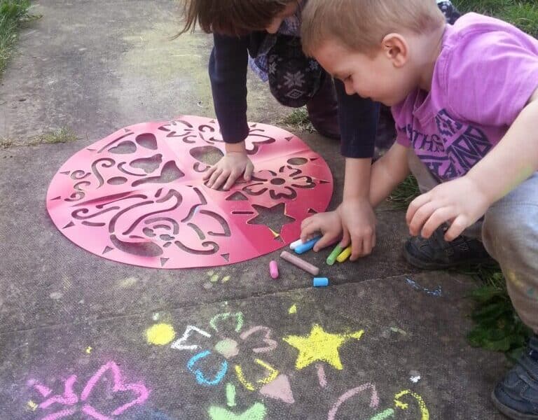 Two young children crouch on a paved path, using coloured chalk and a large red stencil to draw vibrant shapes and flowers on the ground. Colourful chalk drawings and patterns fill the pavement around their hands, with grass visible at the edges.