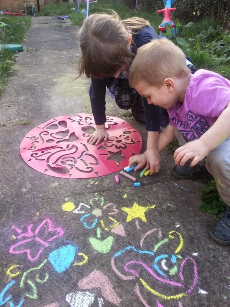 Two young children kneel on a paved path, using colourful chalk and a red stencil to draw shapes and patterns on the ground. Bright chalk drawings of flowers, stars, and other designs are visible on the pavement around them.