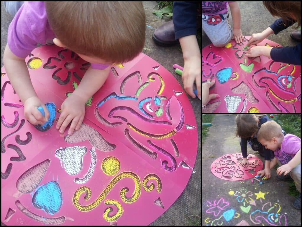 Three young children use coloured chalk and a large pink stencil to create art on pavement. The stencil has various shapes and patterns, and the kids fill them in with blue, yellow, green, and pink chalk, working closely together outdoors.