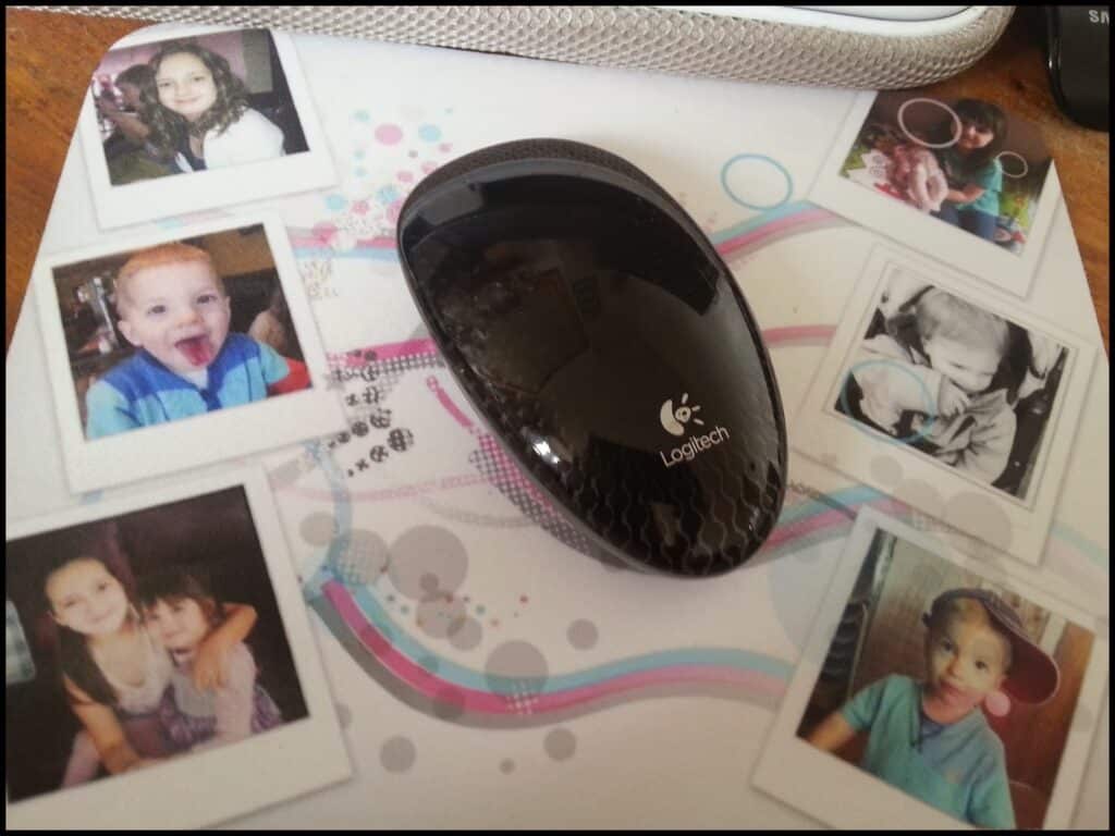 A black Logitech computer mouse sits on a white mouse mat decorated with six Polaroid-style photos of children in various poses, some smiling and some playing, surrounded by colourful swirls and dots. A silver object is partly visible at the top.