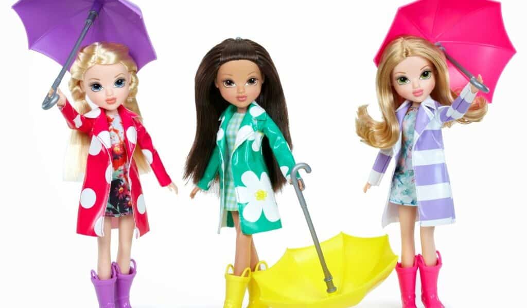 Three fashion dolls stand side by side, each wearing colourful raincoats and boots. The doll on the left has a purple umbrella, the middle doll holds a closed yellow umbrella, and the right doll has a pink umbrella. All three have long hair and floral outfits.