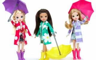Three fashion dolls stand side by side, each wearing colourful raincoats and boots. The doll on the left has a purple umbrella, the middle doll holds a closed yellow umbrella, and the right doll has a pink umbrella. All three have long hair and floral outfits.