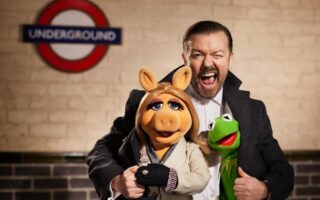 A man in a dark coat smiles widely whilst standing with Miss Piggy and Kermit the Frog puppets. They pose in front of a brick wall with a London Underground sign visible in the background.