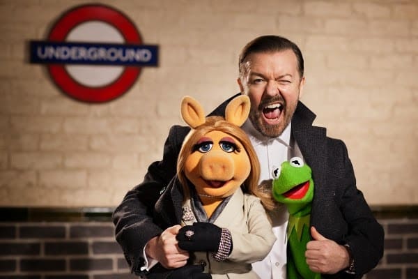 A man in a dark coat smiles widely whilst standing with Miss Piggy and Kermit the Frog puppets. They pose in front of a brick wall with a London Underground sign visible in the background.