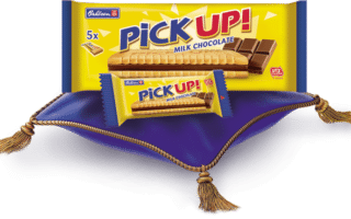 A blue and gold pack of PiCK UP! Milk Chocolate biscuits and a single biscuit bar rest on a royal blue cushion with gold tassels. The packaging displays an image of the rectangular biscuit and chocolate filling. The background is white.