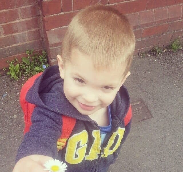 A young boy with light brown hair, wearing a blue hoodie with yellow letters and a red rucksack, stands outside on a pavement by a brick wall, smiling and holding a small white daisy flower towards the camera.