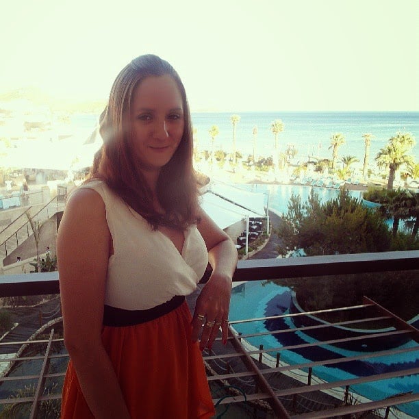 A woman with long brown hair in a white and orange dress stands on a balcony overlooking a bright seaside resort with palm trees, a pool, and the sea in the background under a clear sky. Sunlight softly illuminates part of her face.