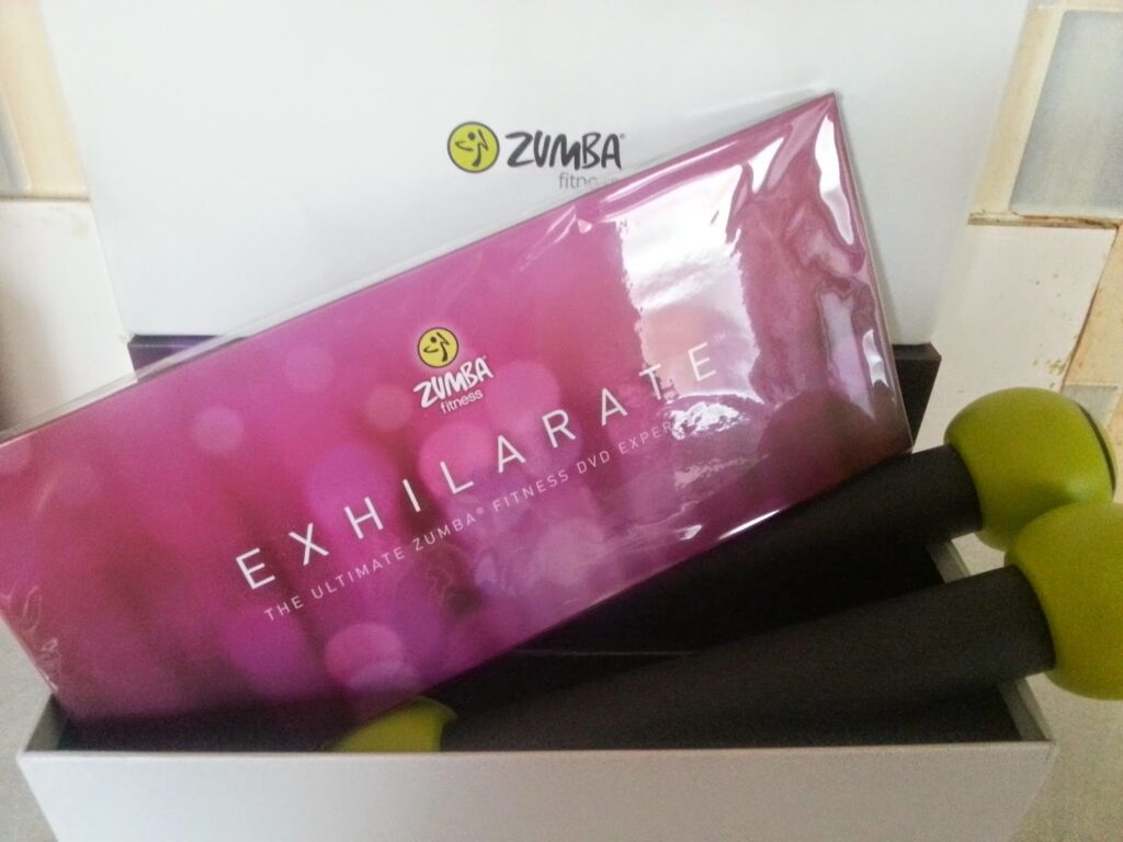 A Zumba Fitness set with a purple "Exhilarate" DVD case, labelled "The Ultimate Zumba Fitness DVD Experience," and two black-and-green Zumba toning sticks inside a white box, set against a light tiled background.
